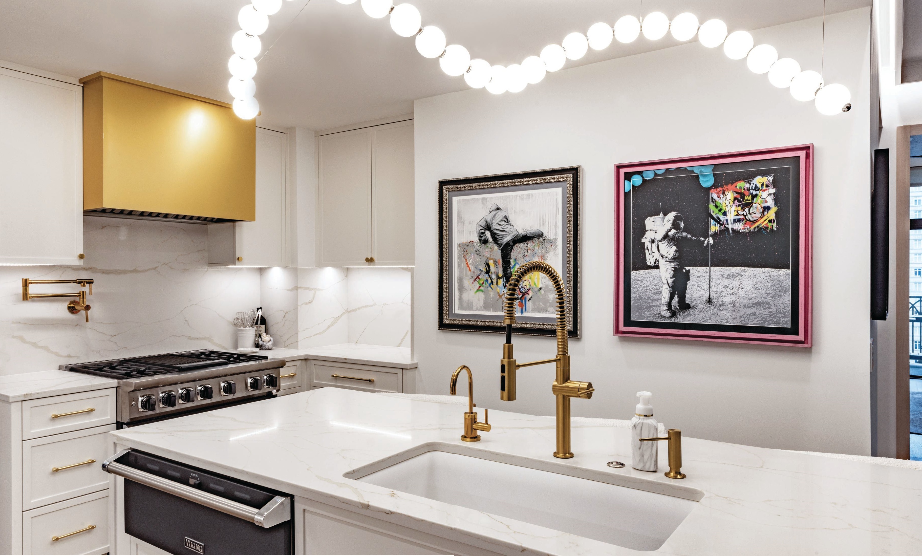 The client’s extensive art collection is featured even in the primary kitchen, where Martin Whatson’s “Climber (Hand Finished)” and “One Small Step (Pink)” add colorful contrast to an Akoya linear chandelier and Difiniti quartz countertops in Pietrasanta Gold. Photographed by Kyle Flubacker
