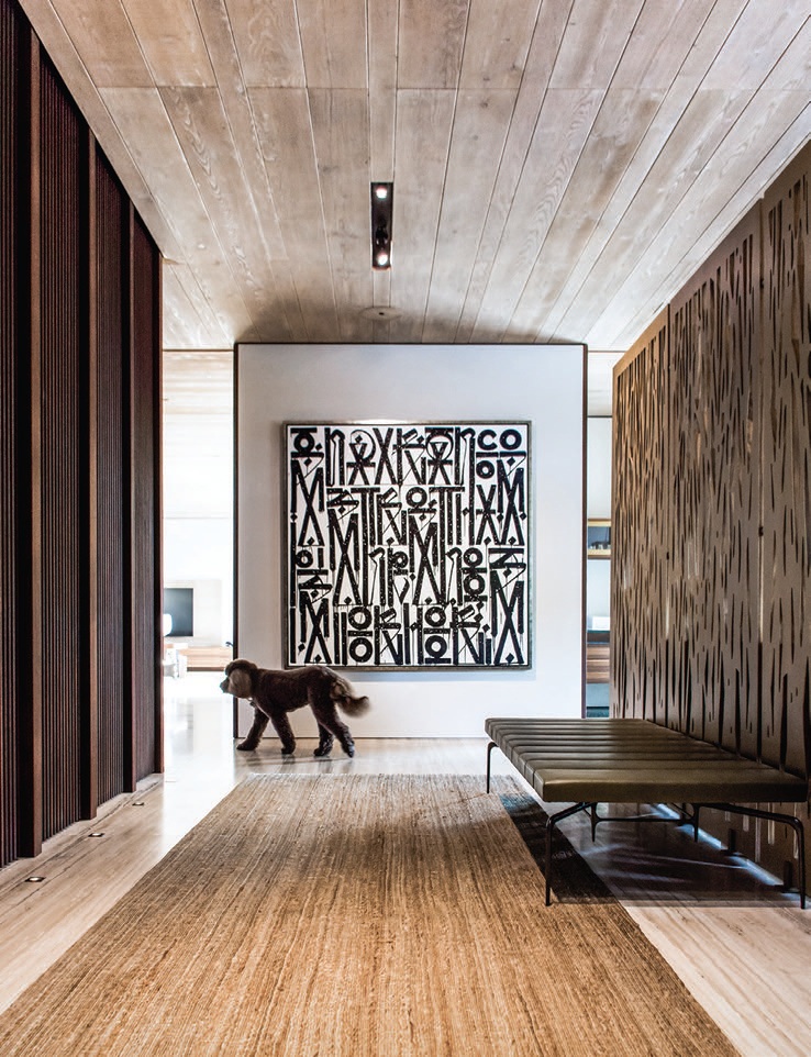 A sisal rug, whose natural texture echoes the striations in the oak ceiling and travertine floor, spans the length of the entryway. The graphic painting is by RETNA PHOTOGRAPHED BY ADRIANA HAMUI